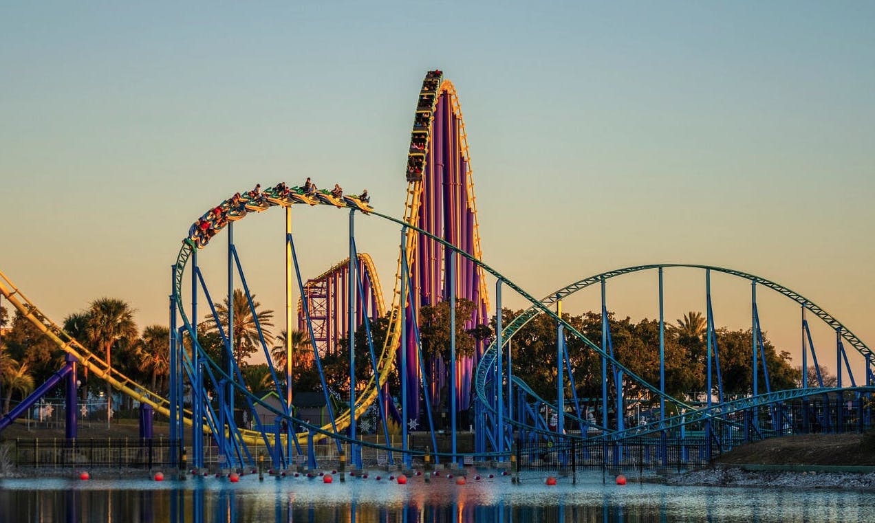 Άνθρωποι που οδηγούν ένα τρενάκι του λούνα παρκ με μπλε και κίτρινες γραμμές, που περιφέρονται πάνω από ένα σώμα νερού στο ηλιοβασίλεμα με δέντρα στο παρασκήνιο.