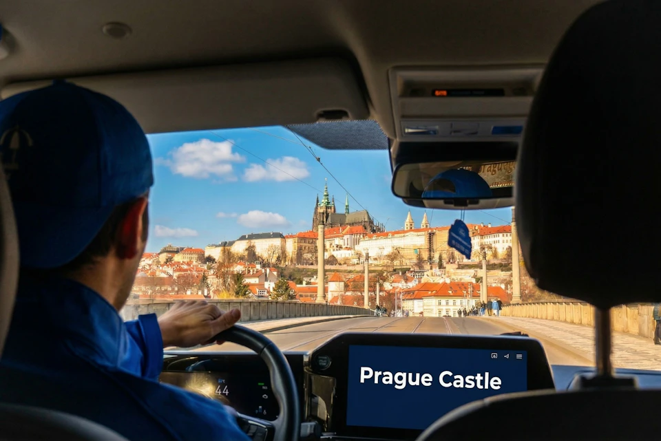 View from inside a car showing Prague Castle in the distance, with the dashboard displaying "Prague Castle."