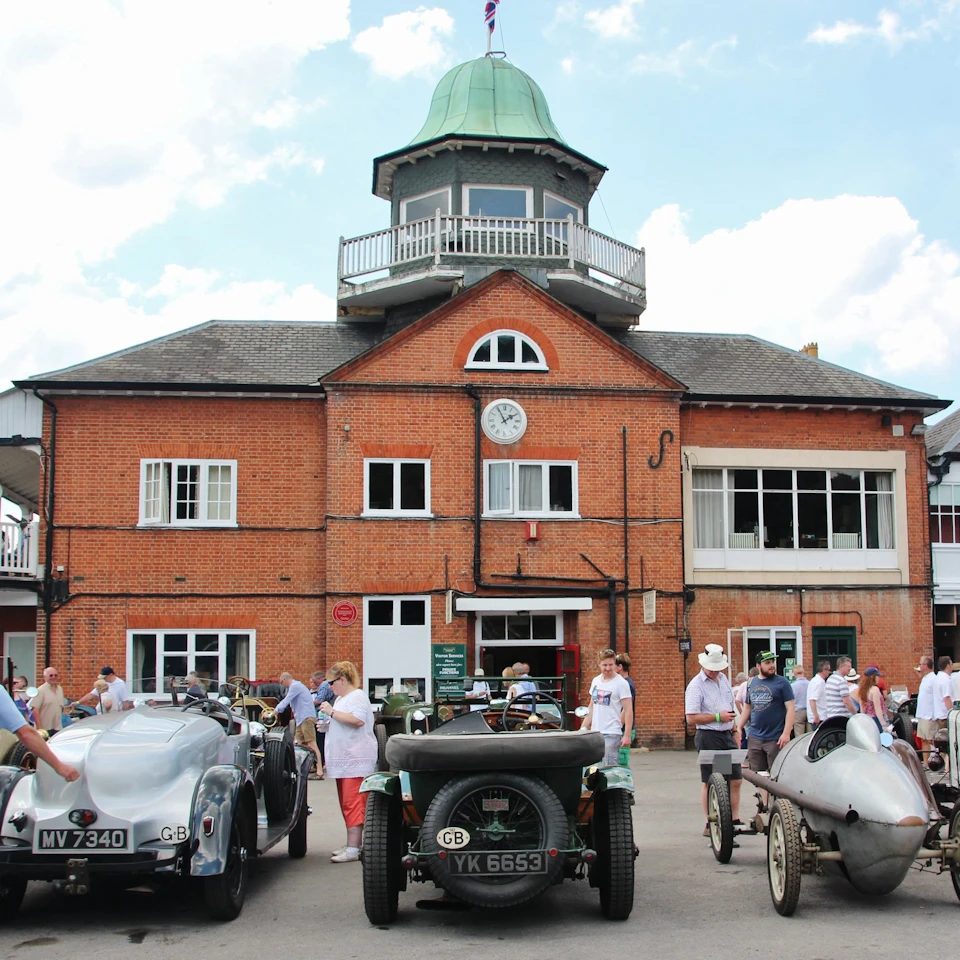 People gather around vintage cars in front of a brick building with a clock and a cupola on the roof.