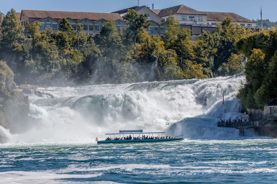 A tour boat with people near a large, cascading waterfall with a building and trees in the background.