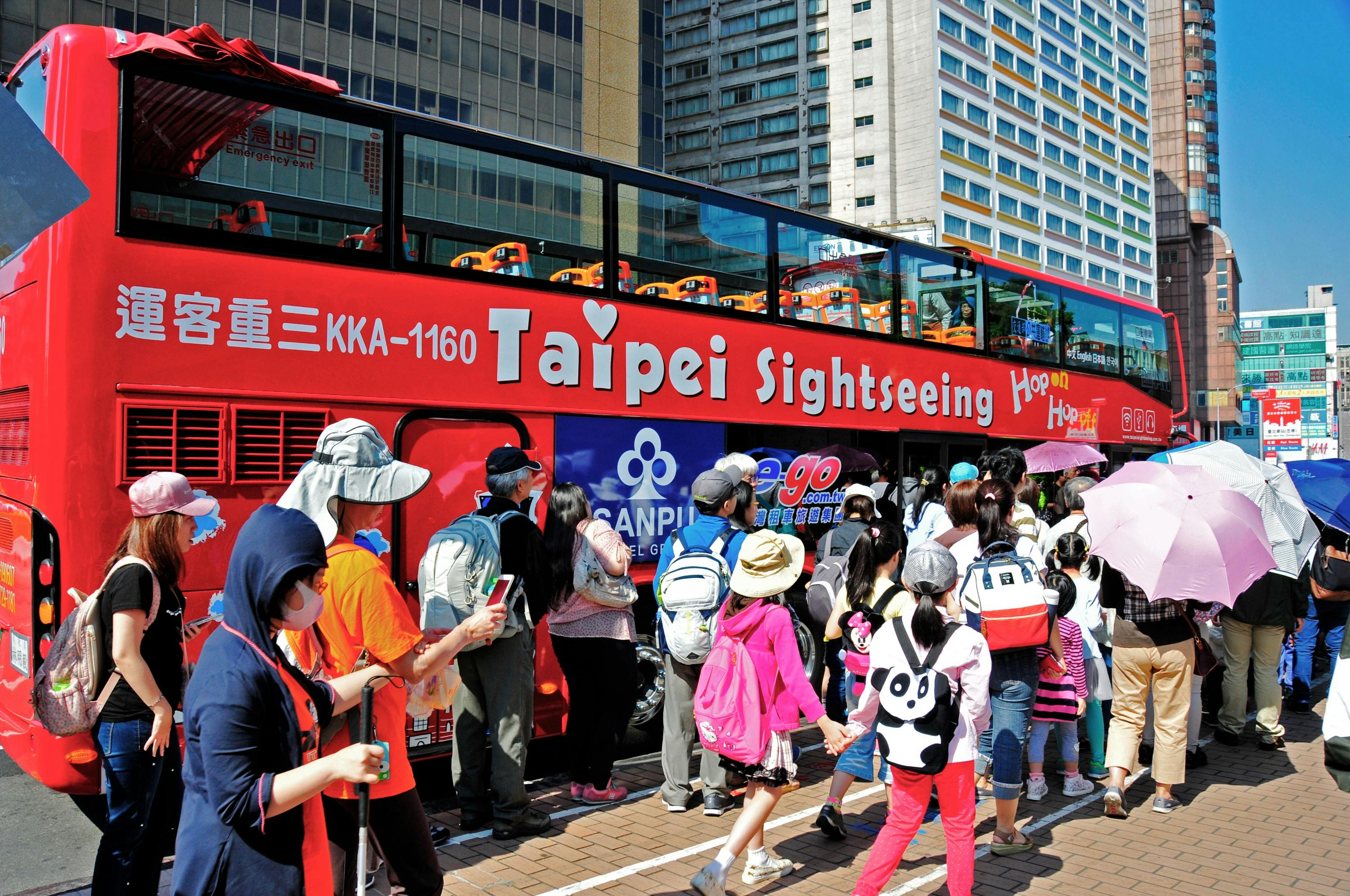 A crowd queues to board a red Taipei Sightseeing double-decker bus in a city street under sunny weather.