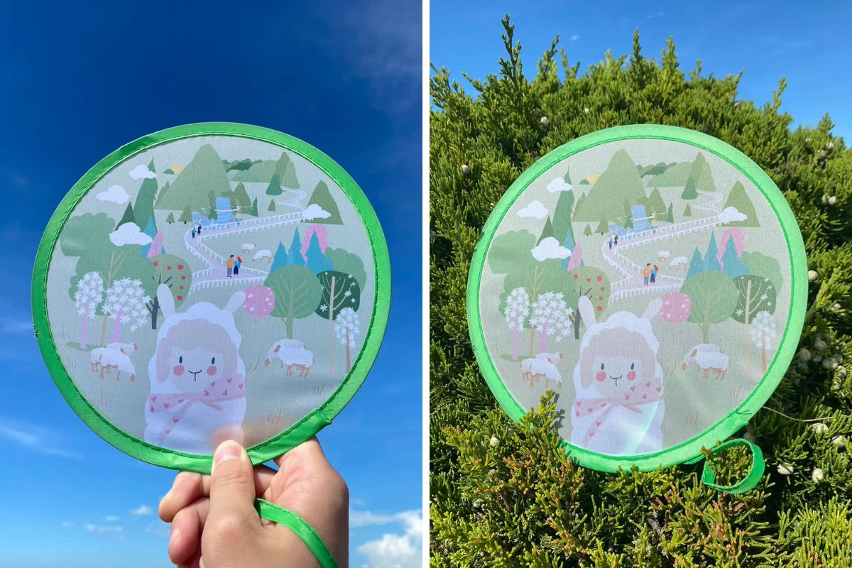 A hand holding a circular fan with a sheep-themed design in front of a blue sky, and in front of bushes.