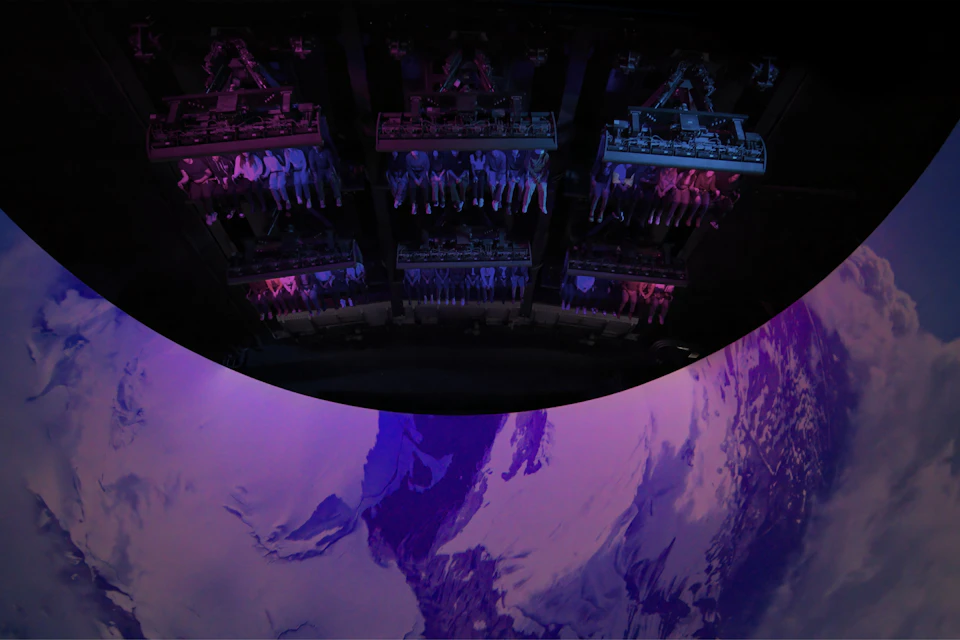 People in a suspended ride above a large curved screen displaying a purple and white mountainous scene.
