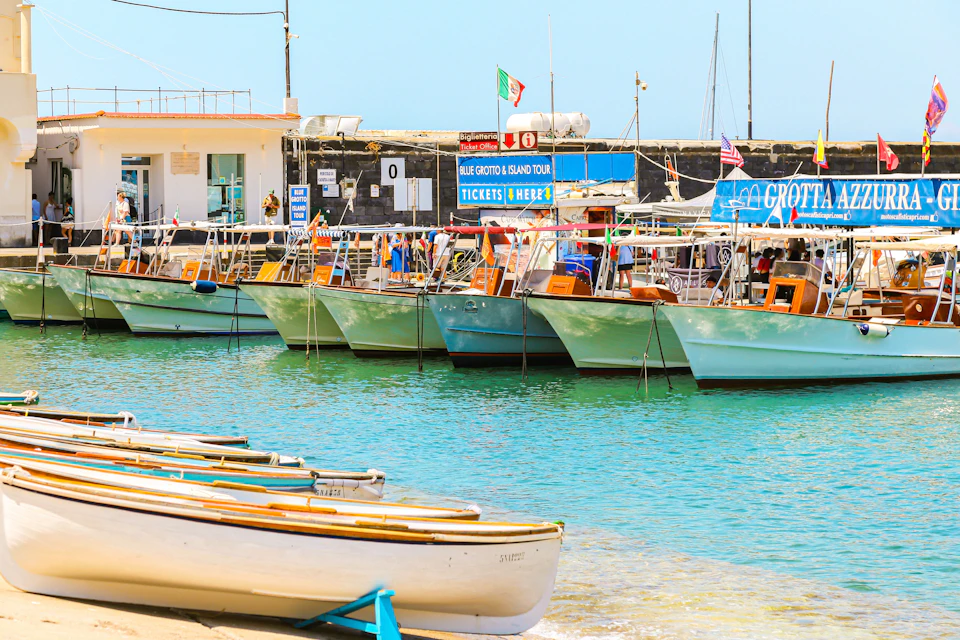 Small boats docked by a pier with colourful signs advertising tours, against a backdrop of clear blue water and sky.