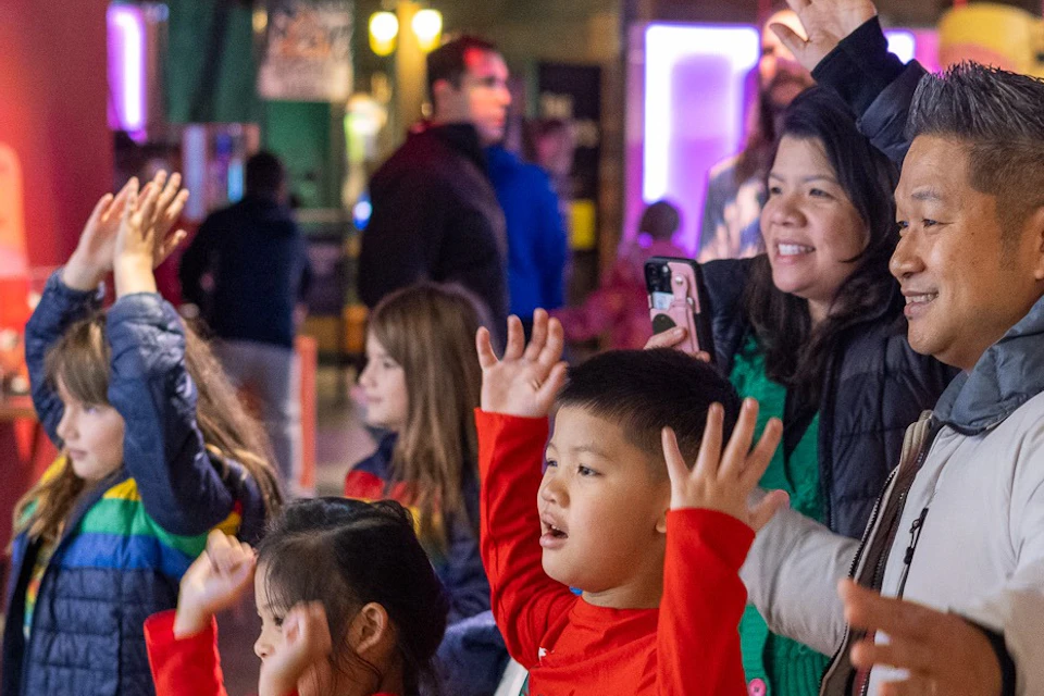 A group of adults and children raising their arms in an interactive exhibit in a brightly lit museum space.
