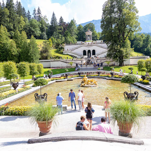 Menschen gehen in der Nähe eines Brunnens mit einer goldenen Statue in einem angelegten Garten spazieren, der zu einem weißen Gebäude mit Stufen und Grünanlagen führt.