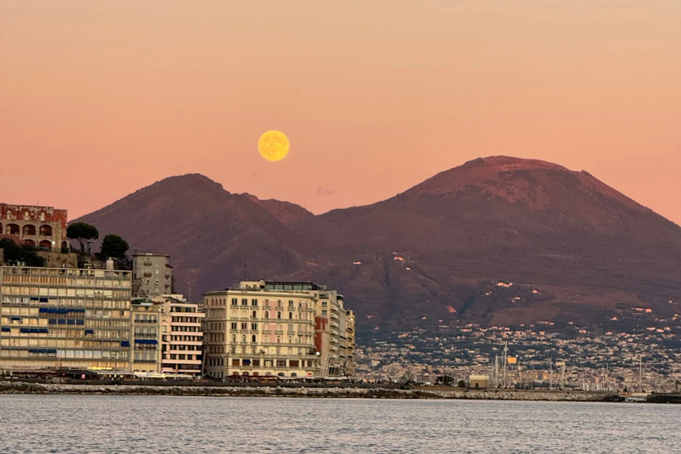 A cityscape by the water at sunset, featuring mountains in the background and a full moon in the sky.