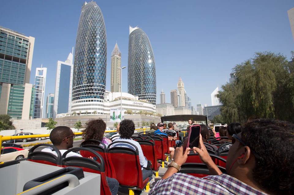Tourists seated on an open-top bus tour, with modern skyscrapers and iconic buildings in the background under a clear blue sky.