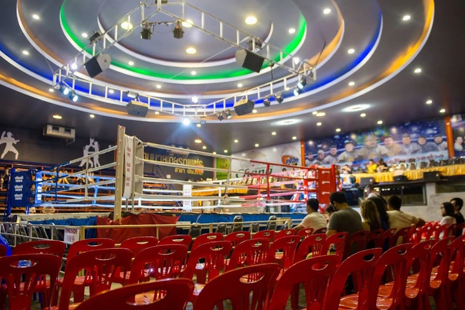 A boxing ring in a brightly lit arena with red plastic chairs in the foreground and colorful banners in the background.