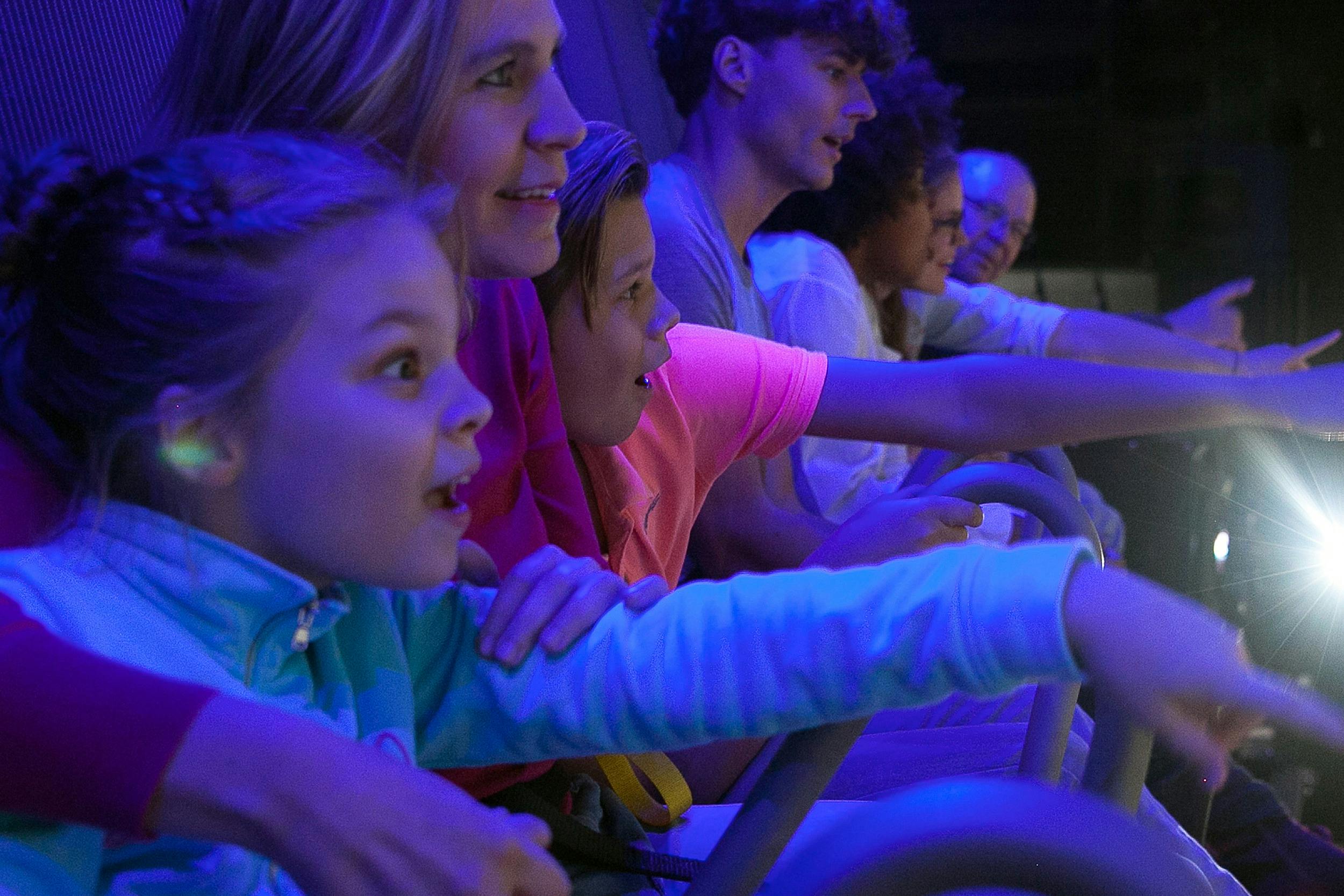 Lidé různého věku něco pozorně sledují, osvětlené fialovým osvětlením. Někteří vyjadřují úctu nebo vzrušení.