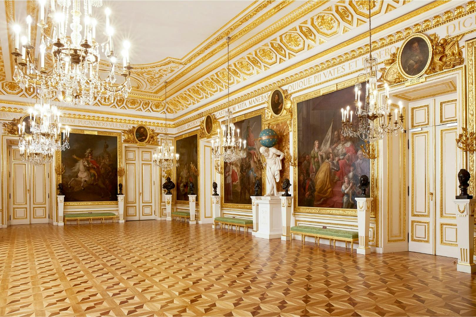 Ein luxuriöser Raum mit kunstvollen goldenen Verzierungen, Kronleuchtern, großen Gemälden und einem Holzparkett.