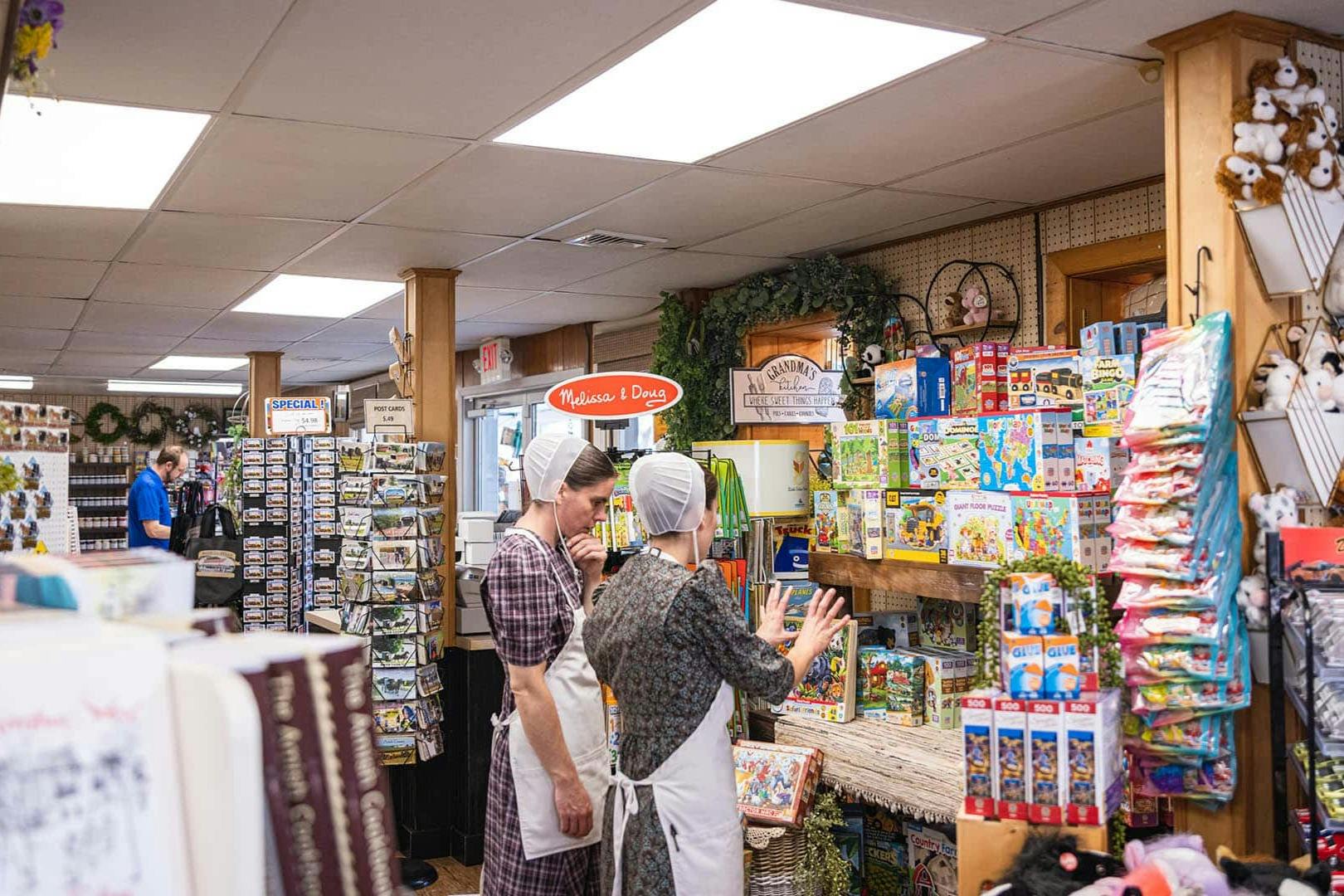 Two women in traditional clothing browse shelves of toys and puzzles inside a well-lit store.