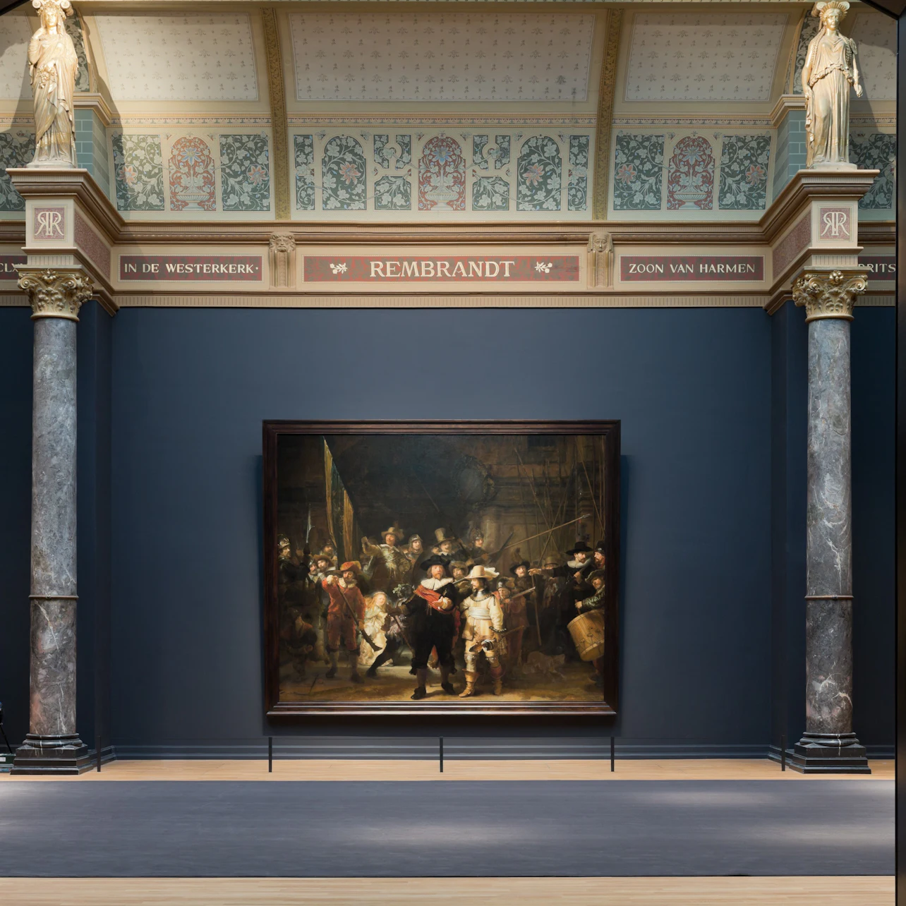 A museum gallery displaying a large painting by Rembrandt, framed by ornate architecture with columns and decorative details.