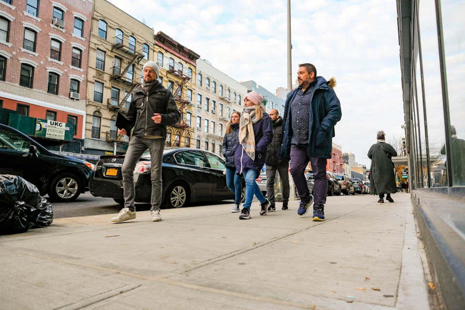 Four people walking on a city sidewalk, dressed in winter clothing, with parked cars and buildings in the background.