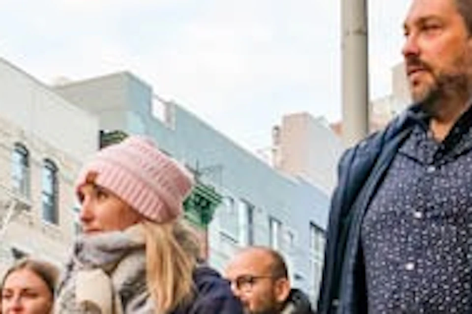 Four people walking on a city sidewalk, dressed in winter clothing, with parked cars and buildings in the background.