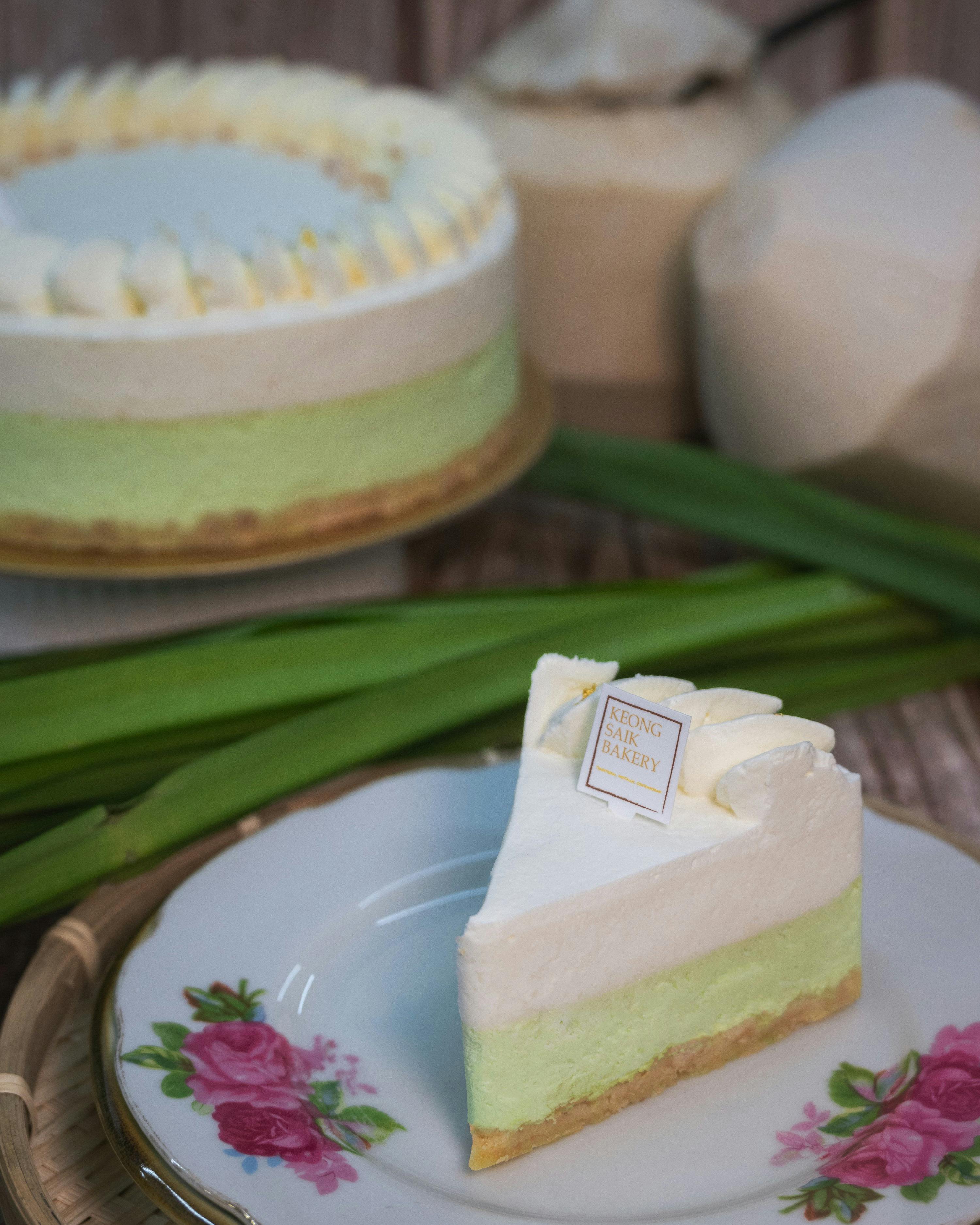 A slice of pandan cake with white frosting on a floral plate, with the whole cake and pandan leaves in the background.