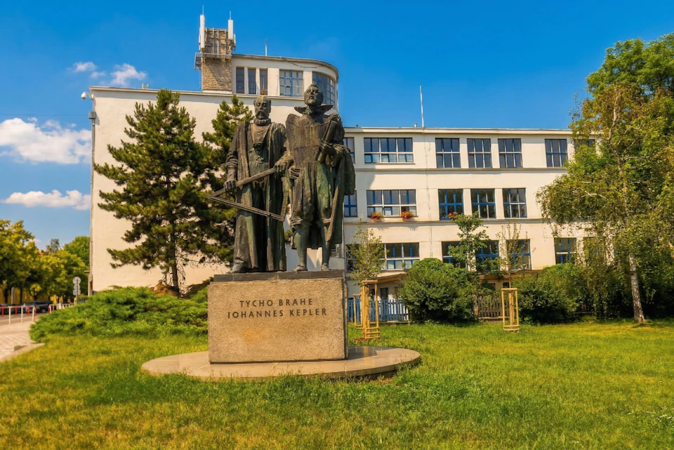 A statue of Tycho Brahe and Johannes Kepler in front of a modern building with trees and grass under a clear blue sky.