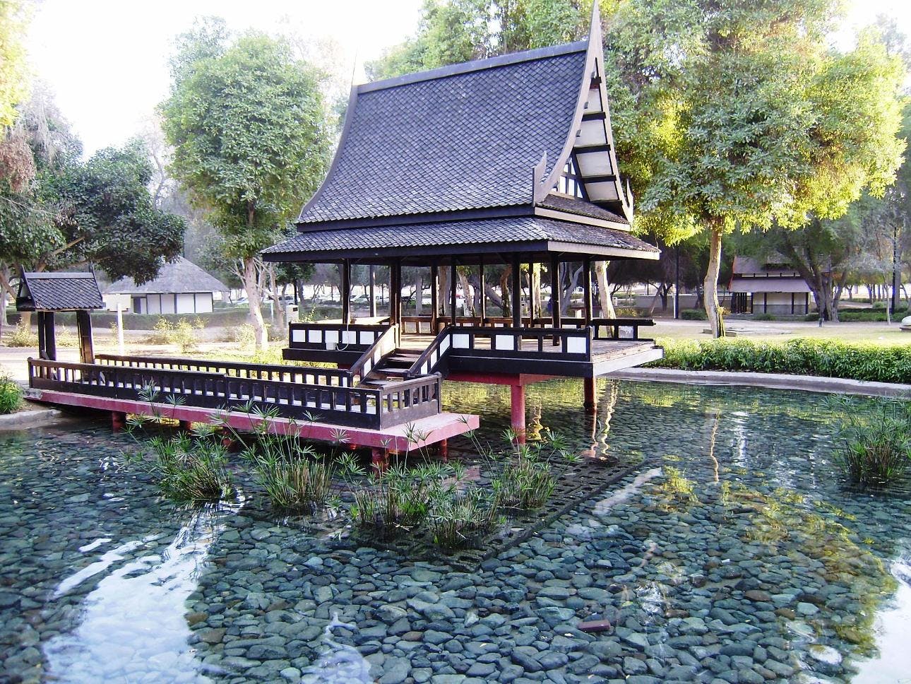 Ένα περίπτερο ταϊλανδέζικου στιλ με μυτερή οροφή στέκεται σε ξυλοπόδαρα πάνω από μια καθαρή λίμνη, που περιβάλλεται από δέντρα και πράσινο.