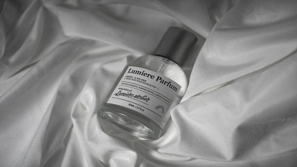 A bottle of Lumiere Parfum placed on a white satin fabric, featuring a silver cap and a minimalist label design.