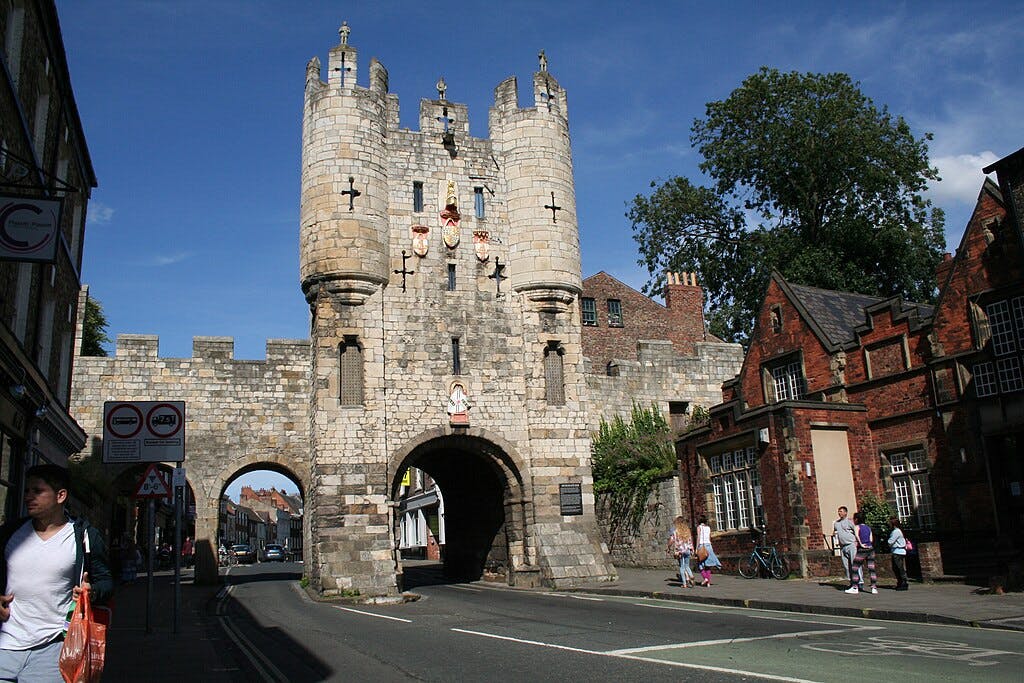 Kamienna strażnica z dwiema wieżami w zabytkowym mieście, otoczona murowanymi budynkami i kilkoma pieszymi na ulicy.