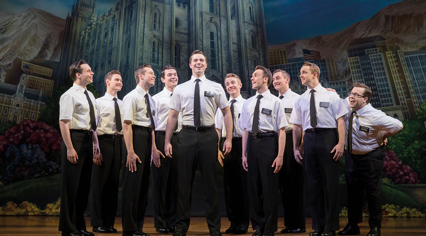 A group of men in white shirts, black pants, and ties stand smiling on stage against a backdrop resembling a grand building.