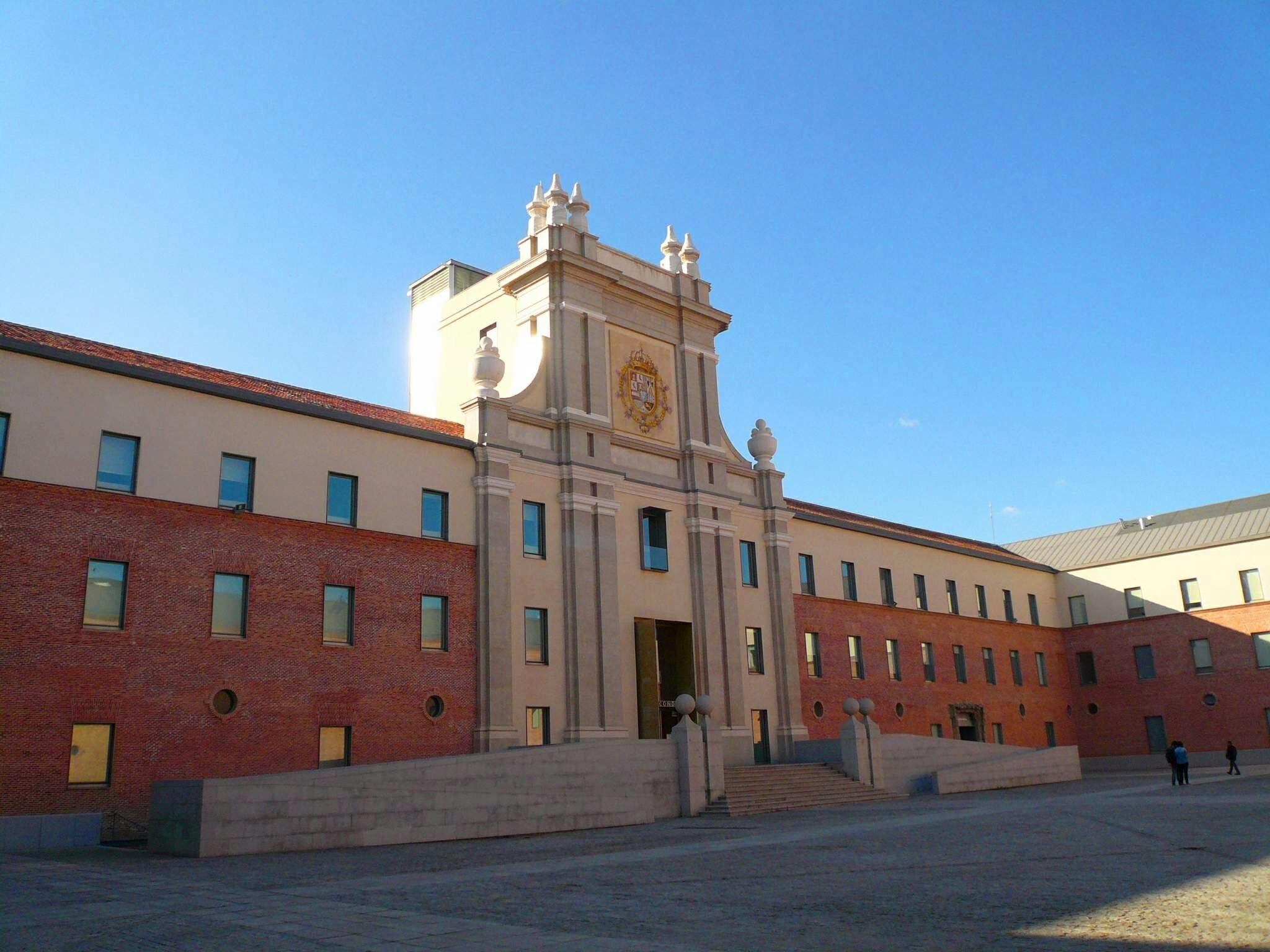 一栋大型砖石建筑，窗户对称，中央塔楼坐落在晴朗的蓝天下。 庭院里很少有人。