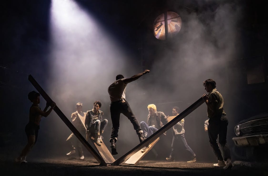 En person balancerer på en skrå platform, mens fire andre ser på under dramatisk belysning i en røgfyldt atmosfære.