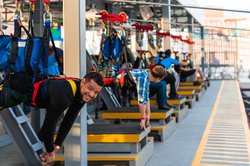 People harnessed and ready for a zipline ride, with one man smiling at the camera. The background shows platforms and equipment.