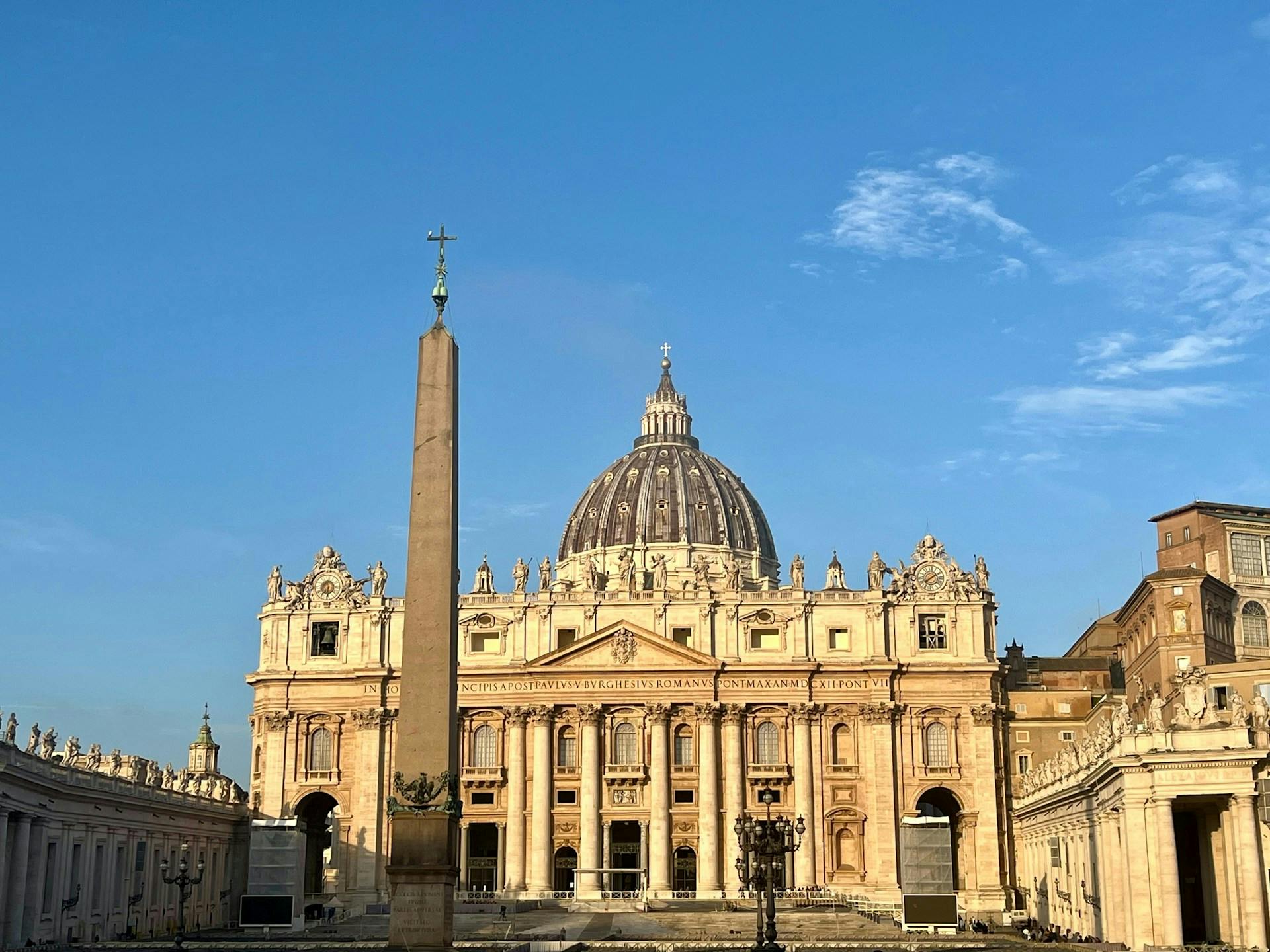 A grand basilica with a large domed roof and ornate facade, fronted by an obelisk, under a clear blue sky.