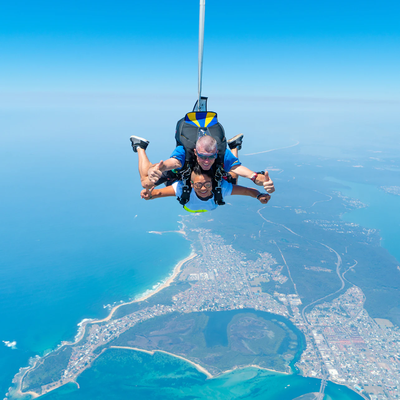 Two people tandem skydiving over a coastal city, with the ocean, beaches, and town visible below against a clear blue sky.