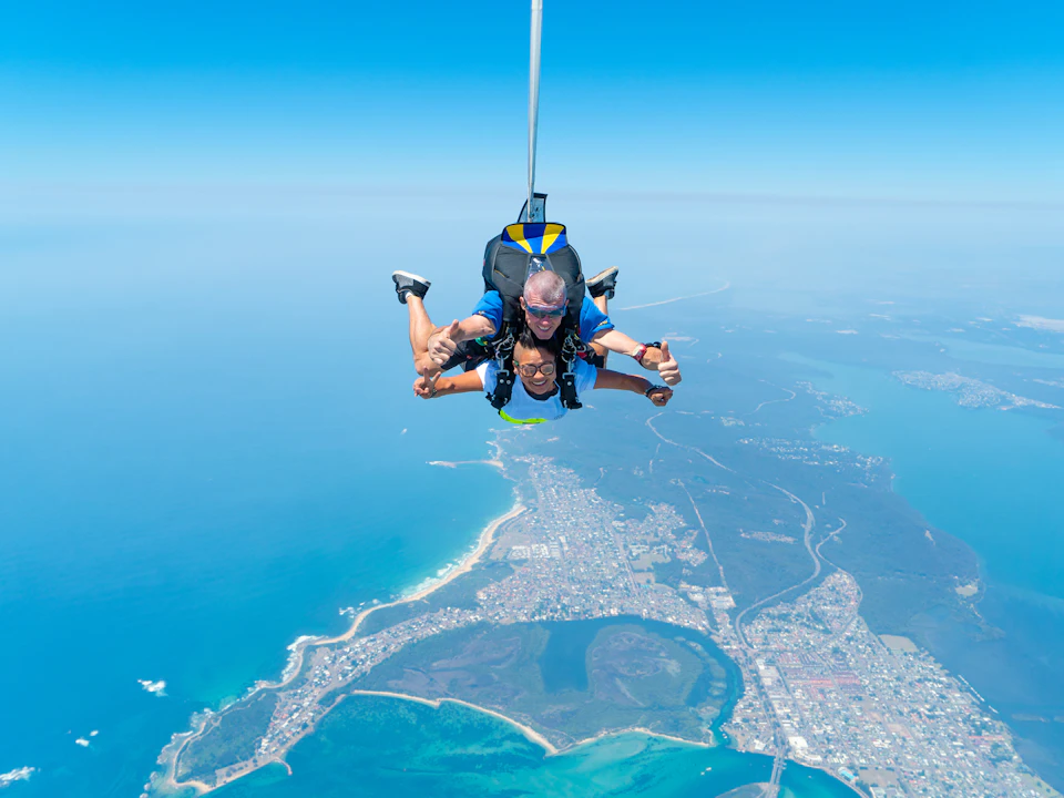 Two people tandem skydiving over a coastal city, with the ocean, beaches, and town visible below against a clear blue sky.