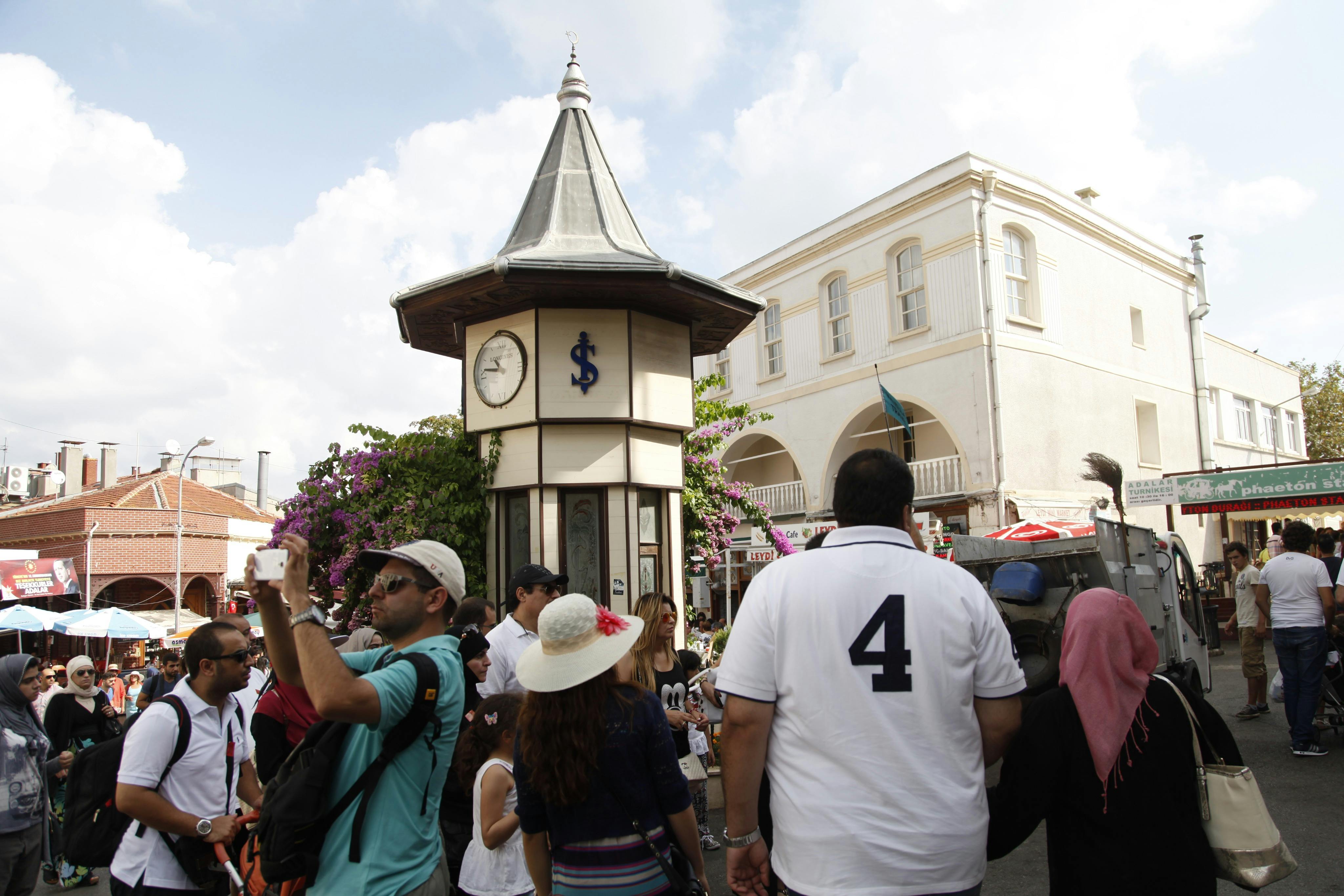 Люди гуляют и фотографируют возле небольшой часовой башни с голубым знаком доллара, под полуоблачным небом.