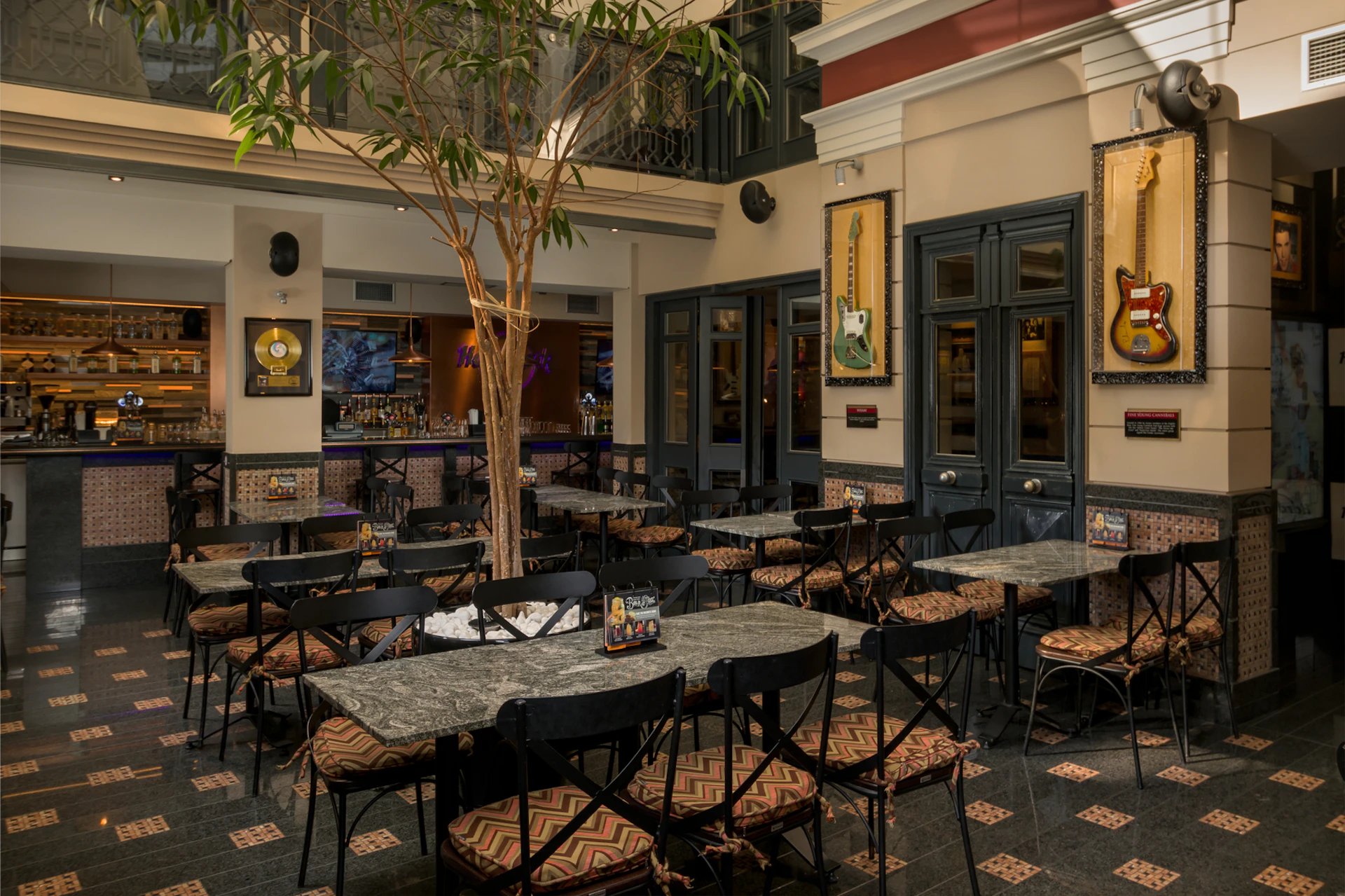 Indoor café with empty tables and chairs, a tree in the center, framed records, and a guitar on the wall. Bar area in the background.