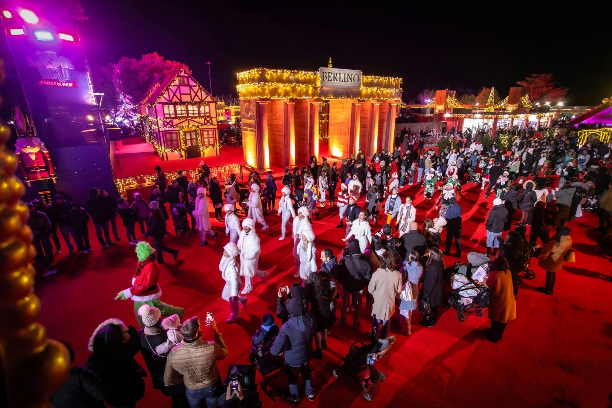A festive nighttime event with crowds, costumed performers, illuminated buildings, and holiday decorations.