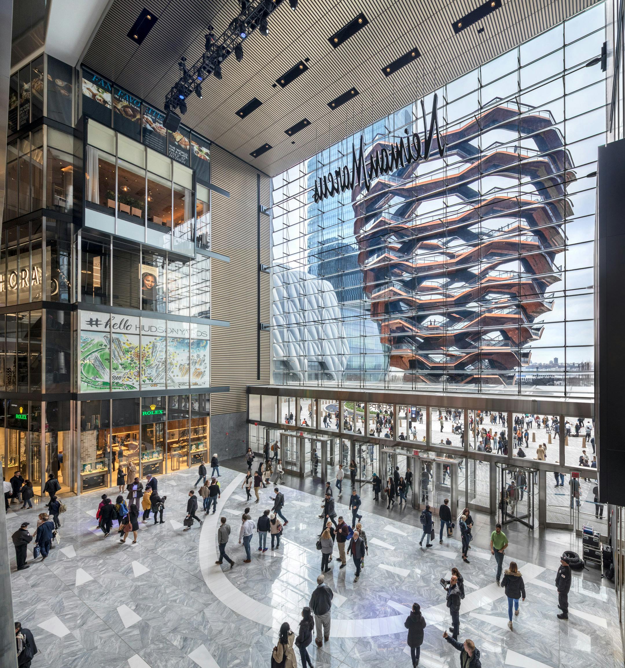 Shoppers in a modern mall with large glass windows and a view of an elaborate, honeycomb-like structure outside.