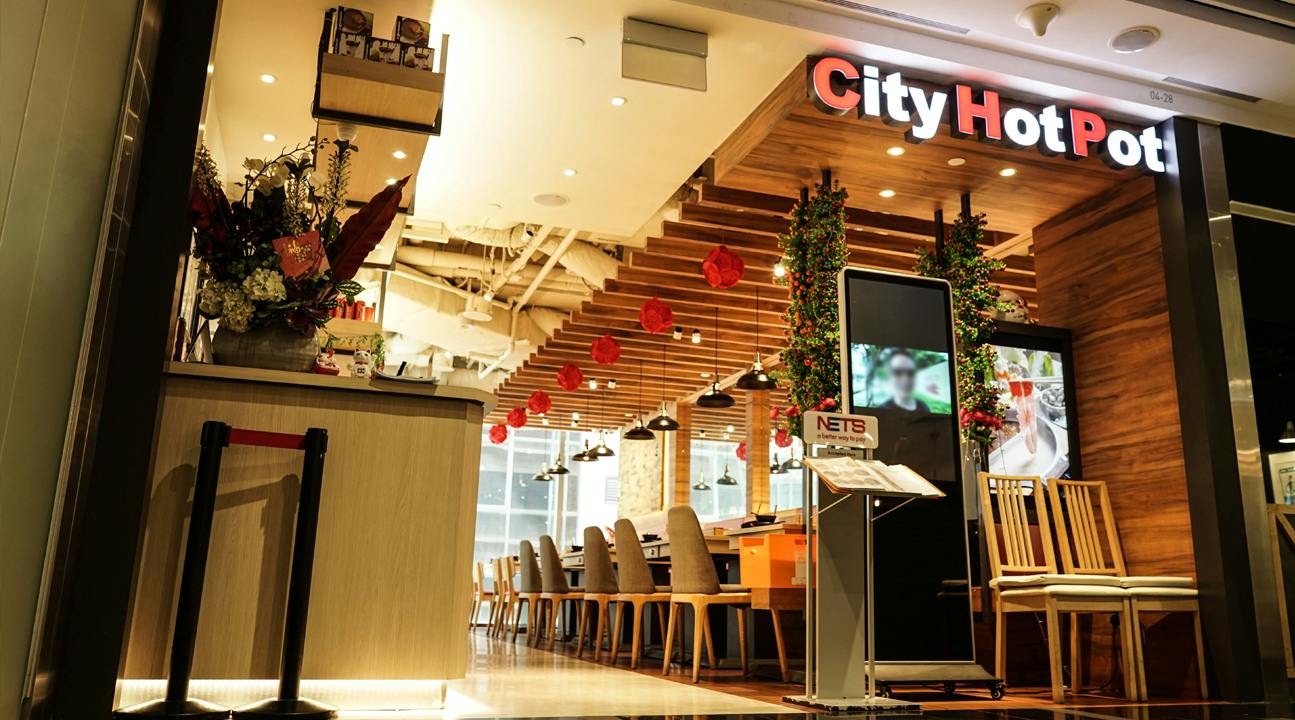 Wejście do restauracji z napisem „City Hot Pot”, ozdobione roślinami i światłami, przedstawiające miejsca siedzące i wyświetlacz menu.
