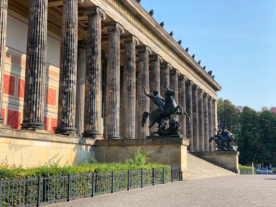 A grand building with tall columns and equestrian statues flanking its entrance steps. Neatly manicured bushes line the foreground.