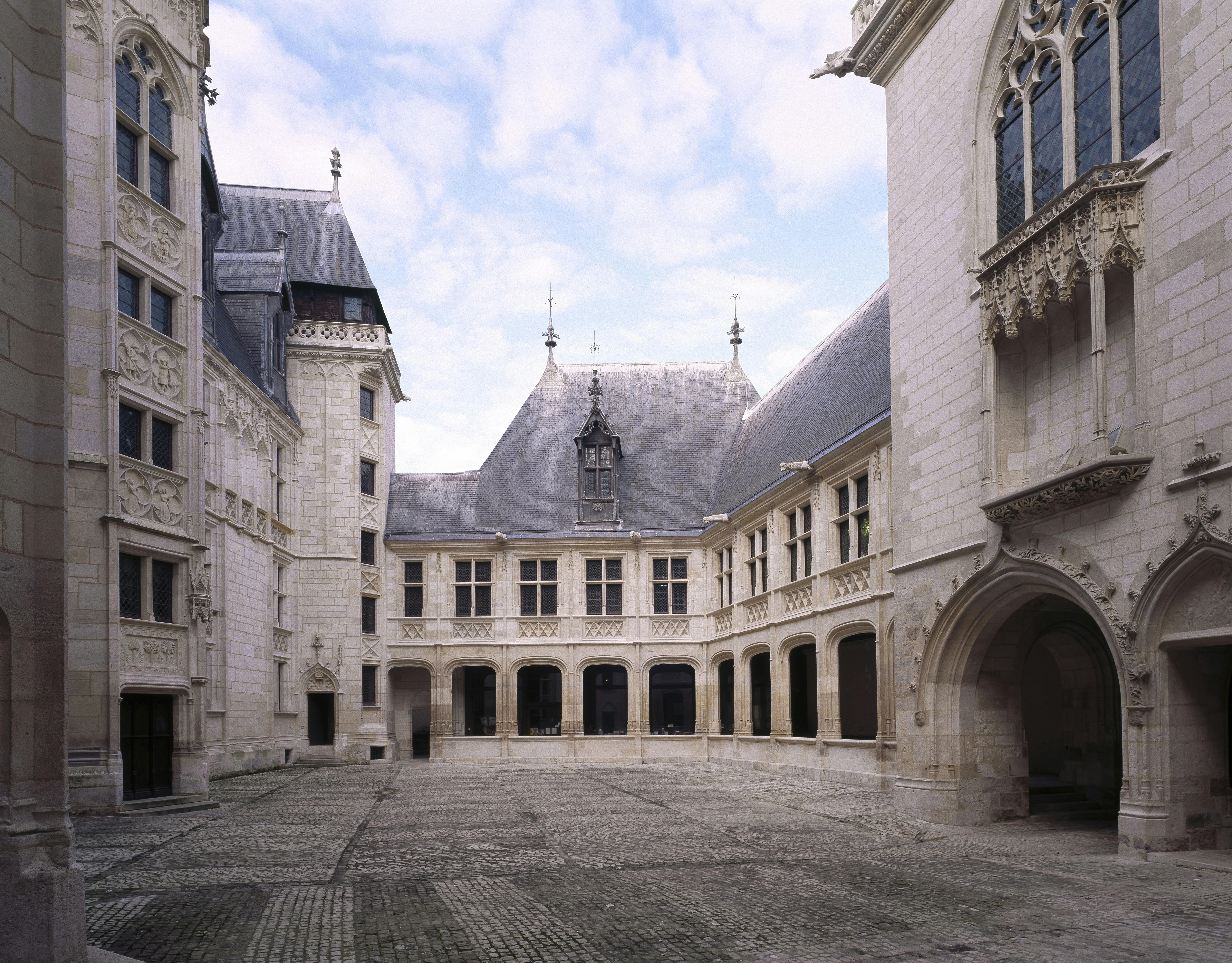 Cour en pierre entourée d'une architecture gothique ornée de plusieurs étages avec des fenêtres cintrées et des détails décoratifs. Ciel nuageux au-dessus.