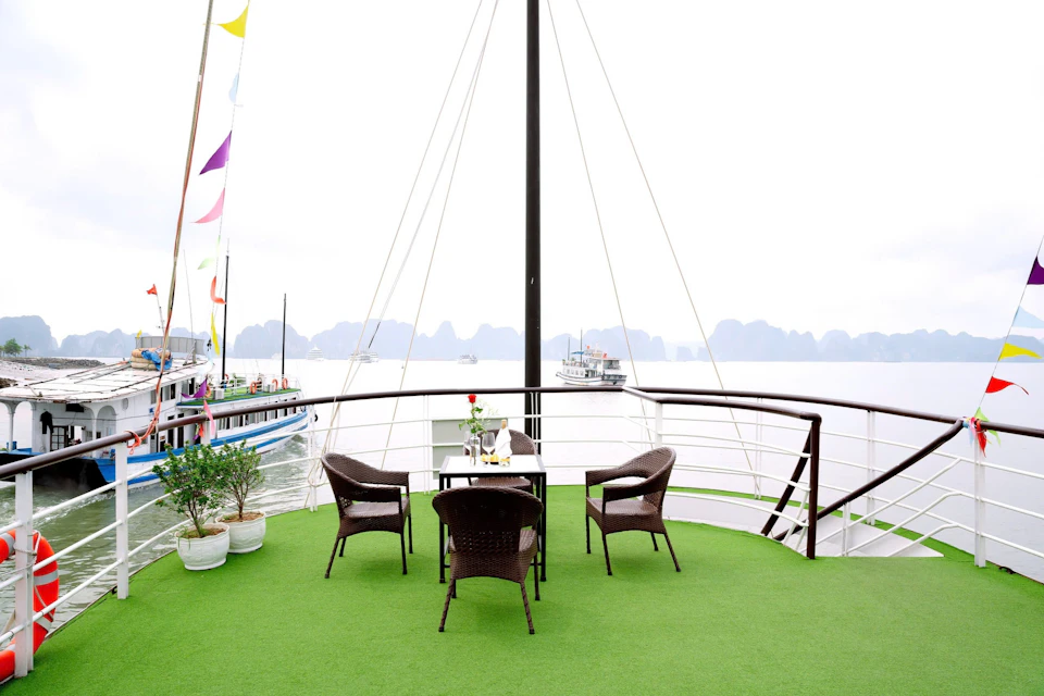 Outdoor deck with green flooring, three brown wicker chairs around a table, potted plants, and boats on water in the background.