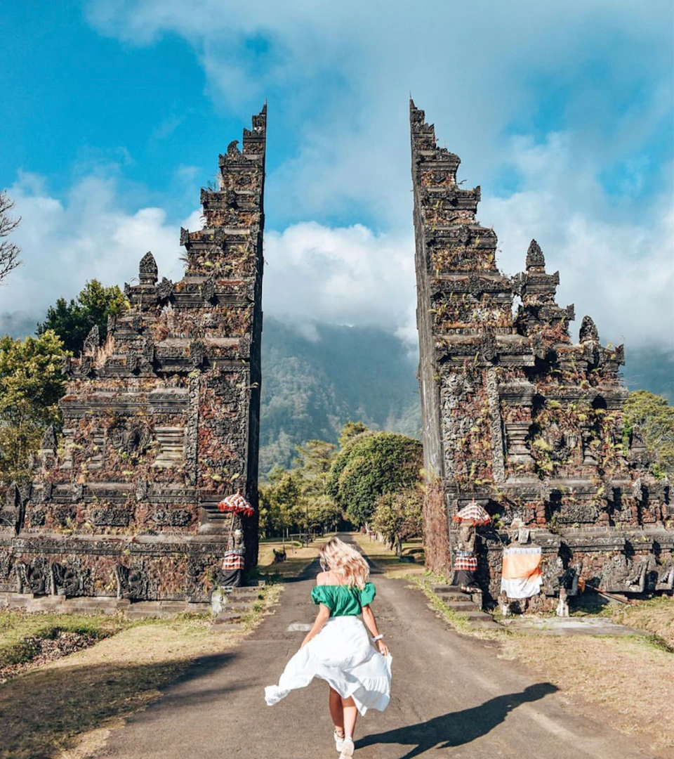 A person in a green shirt and white skirt walks towards an ornate stone gate with mountain and cloudy sky in the background.