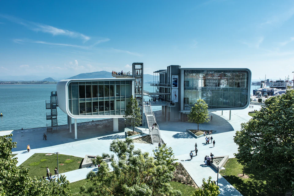 Modern waterfront building with glass façade, an elevated walkway, and outdoor stairs. People walking and trees scattered around.