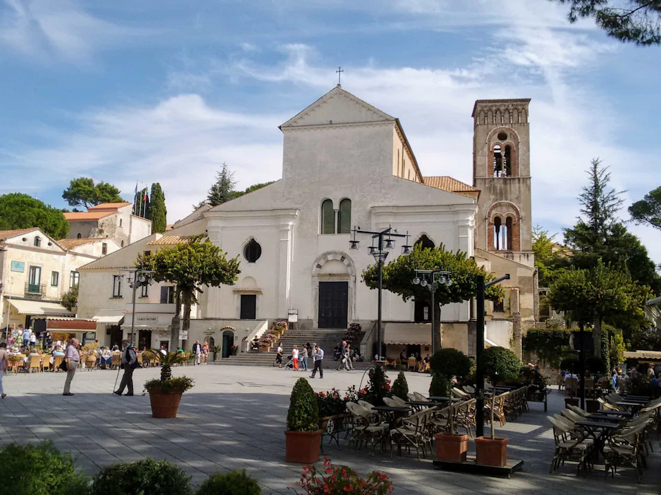 People walking and sitting on a large courtyard with potted trees and chairs, in front of a historic church and bell tower.