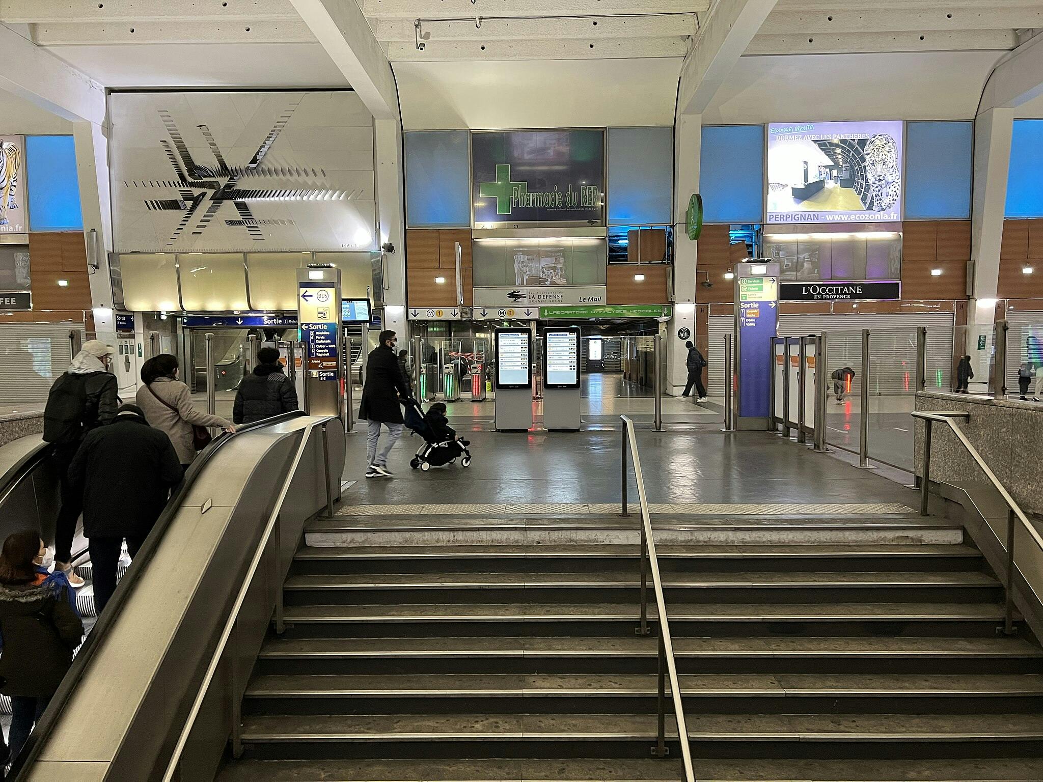 Persones en una moderna estació de tren que es mouen cap a les escales mecàniques i la senyalització digital, amb botigues i taulers publicitaris de fons.