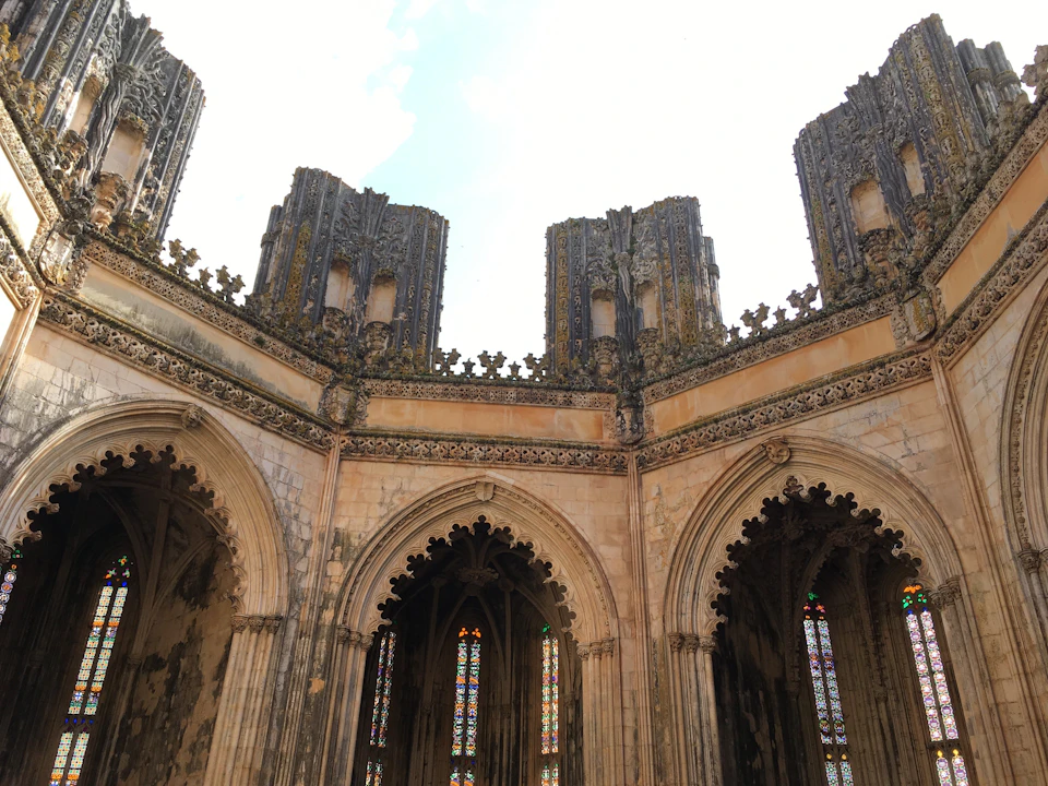 Open-top, gothic-style stone structure with tall, ornate towers and arches featuring stained glass windows below a cloudy sky.