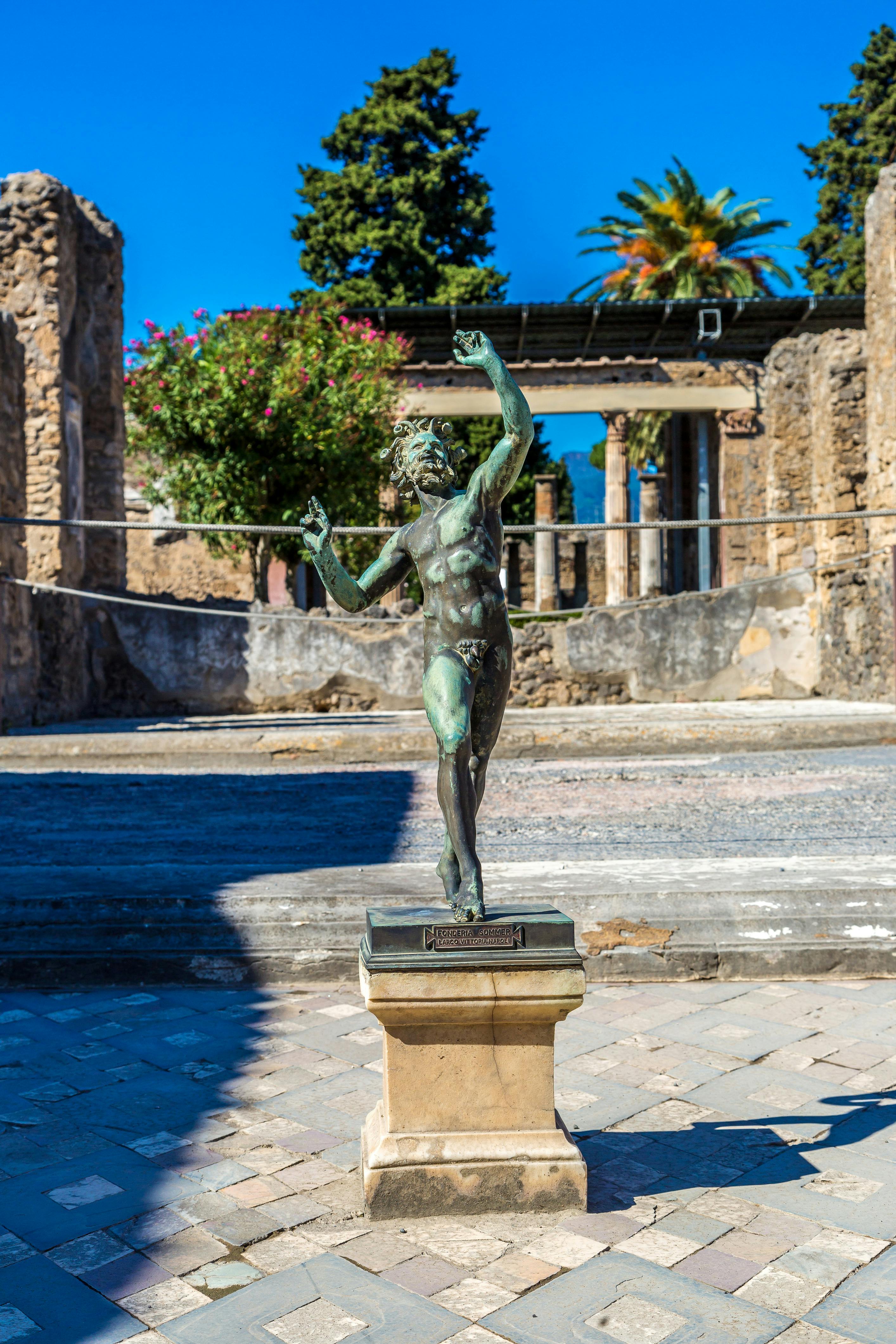 A bronze statue of a dancing figure stands on a pedestal amid ancient ruins with a flowering bush and clear blue sky in the background.