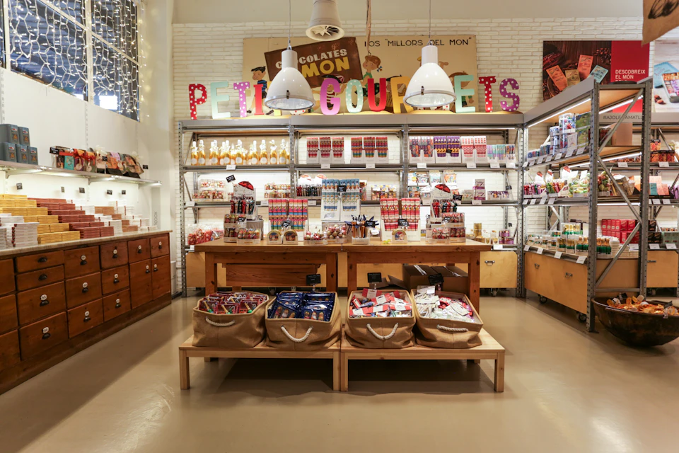 Colorful candy store display with labeled shelves of chocolates and sweets, a wooden table, and large hanging lights overhead.