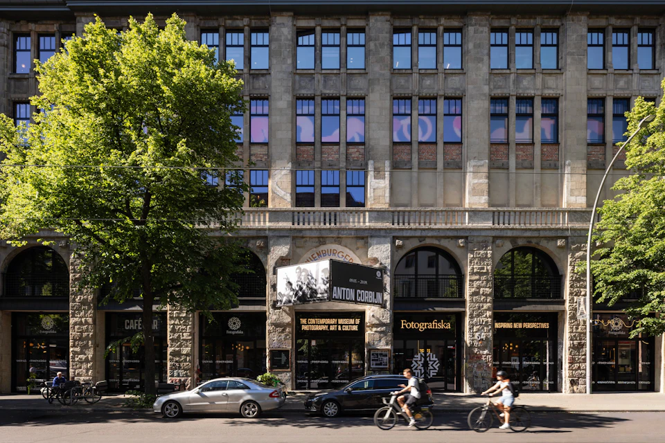 Street view of a building with large windows hosting Fotografiska, with pedestrians, cyclists, and parked cars in front.