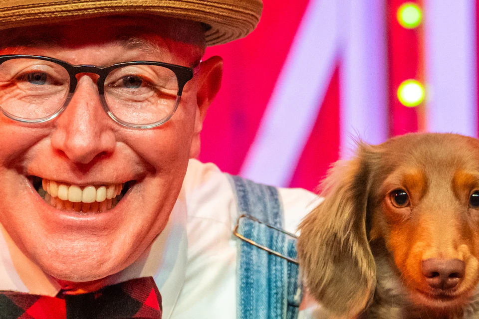 A man in overalls and a bow tie holds a small dog in each hand, standing in a colorful setting with a red and white backdrop.