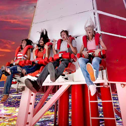 Four people in red harnesses on a thrill ride, showing expressions of excitement and fear, with a city skyline below at sunset.