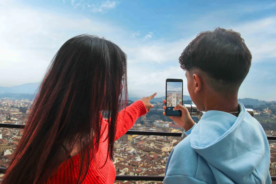 Two people standing on a balcony, with one pointing and the other taking a photo of a cityscape with a smartphone.